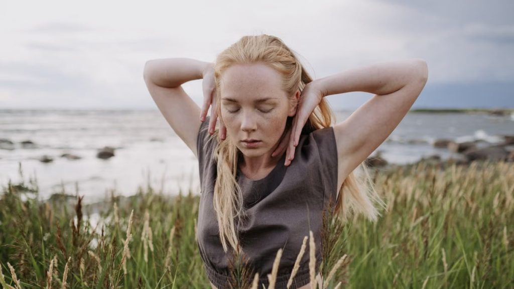 A woman gracefully dances with closed eyes in a coastal grass field, embracing nature.